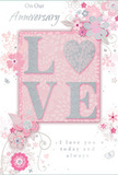 The anniversary card in this image carries a mood of soft, enduring affection, its design as calm and romantic as a quiet evening near the tree-lined streets of Swiss Cottage. The card's surface is covered in a gentle blush-pink tone, smooth and even, creating a tranquil backdrop for the ornate central motif. Prominently placed is the word LOVE, rendered in sizeable, raised letters that are lightly dusted with glitter along their edges. The glitter has a silvery, pearly quality, catching light in a subdued way that makes each letter appear softly luminous, like petals edged with frost. Surrounding this central word, a cluster of stylised flowers, slender leaves, and curling vines spreads outward in graceful arcs. The linework is delicate-pale grey, white, and hints of silver foil-suggesting the fluid sketch of a florist imagining an anniversary bouquet destined for a cosy flat off Avenue Road or a home near the Jubilee line. The blossoms are abstract yet recognisably floral, their overlapping petals and stems forming an airy, circular frame around the central message. Here and there, tiny decorative elements-whorls, dots, and hints of butterfly-like shapes-add whimsy and softness, preventing the design from feeling static. Inside, part of the printed message is visible in elegant script, relaying affectionate, lasting sentiments suitable for celebrating years of partnership. Taken together, the blush background, raised glittered letters, and fine floral detailing give the card the character of a paper bouquet-quiet, refined, and deeply sentimental. It feels perfect for presenting alongside fresh flowers from a local Swiss Cottage florist, marking an anniversary with a keepsake that speaks gently but sincerely of love that has stood the test of time.